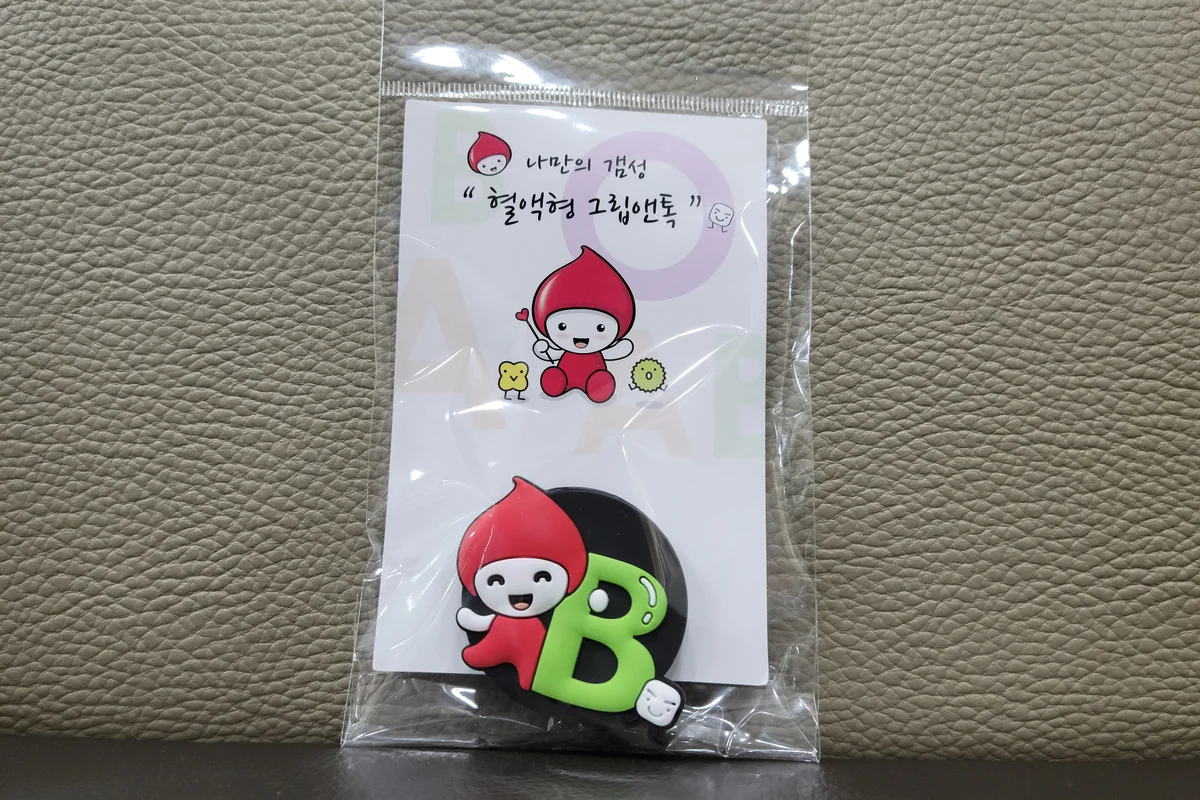 A Korean Red Cross blood-donation souvenir, a "혈액형 그립앤톡" blood-type phone grip: a cartoon blood-drop mascot beside a big green letter B