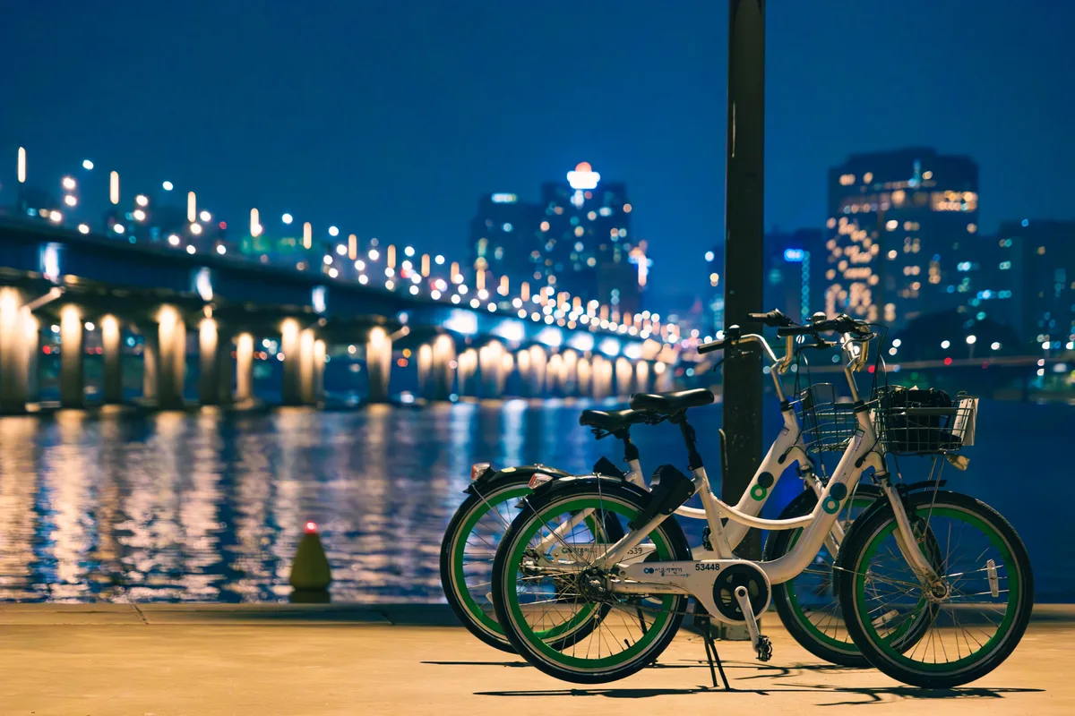 Public share-bikes parked along Seoul's Han River at night, the lit bridge and skyline behind — the kind of after-work scene increasingly replacing the company drinking session