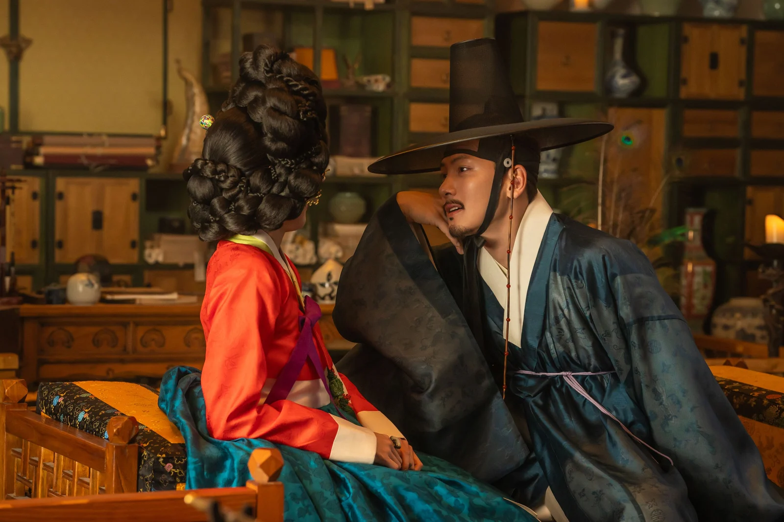 지창욱 as 조원, in a dark blue robe and gat, leaning towards a woman in a red jeogori in a lamplit room
